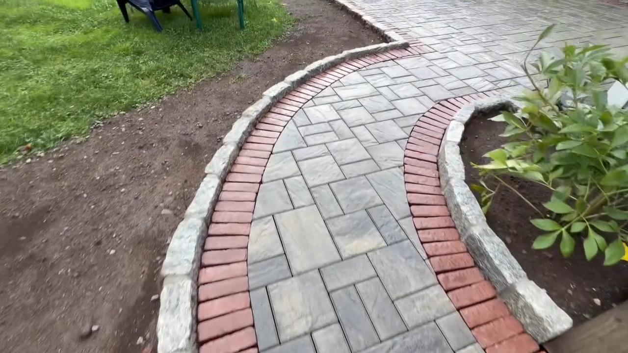 Curved paver walkway with brick border and natural stone edging beside landscaped planting beds